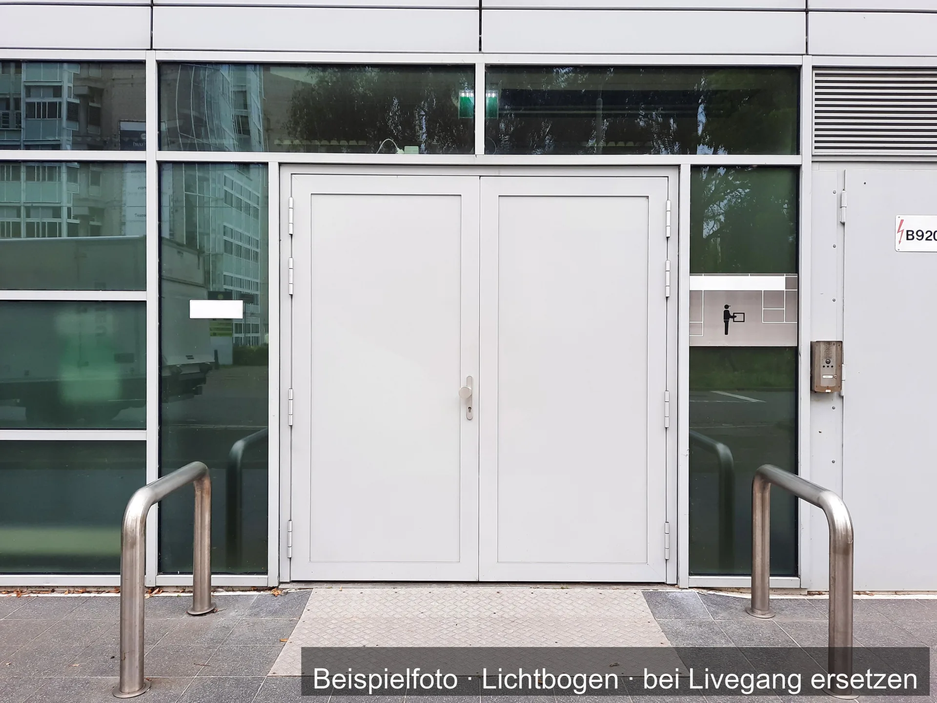 Hellgraue zweiflügelige Eingangstür in einer Fassade aus Glas, davor zwei Bügel aus Metall auf dem Gehweg.