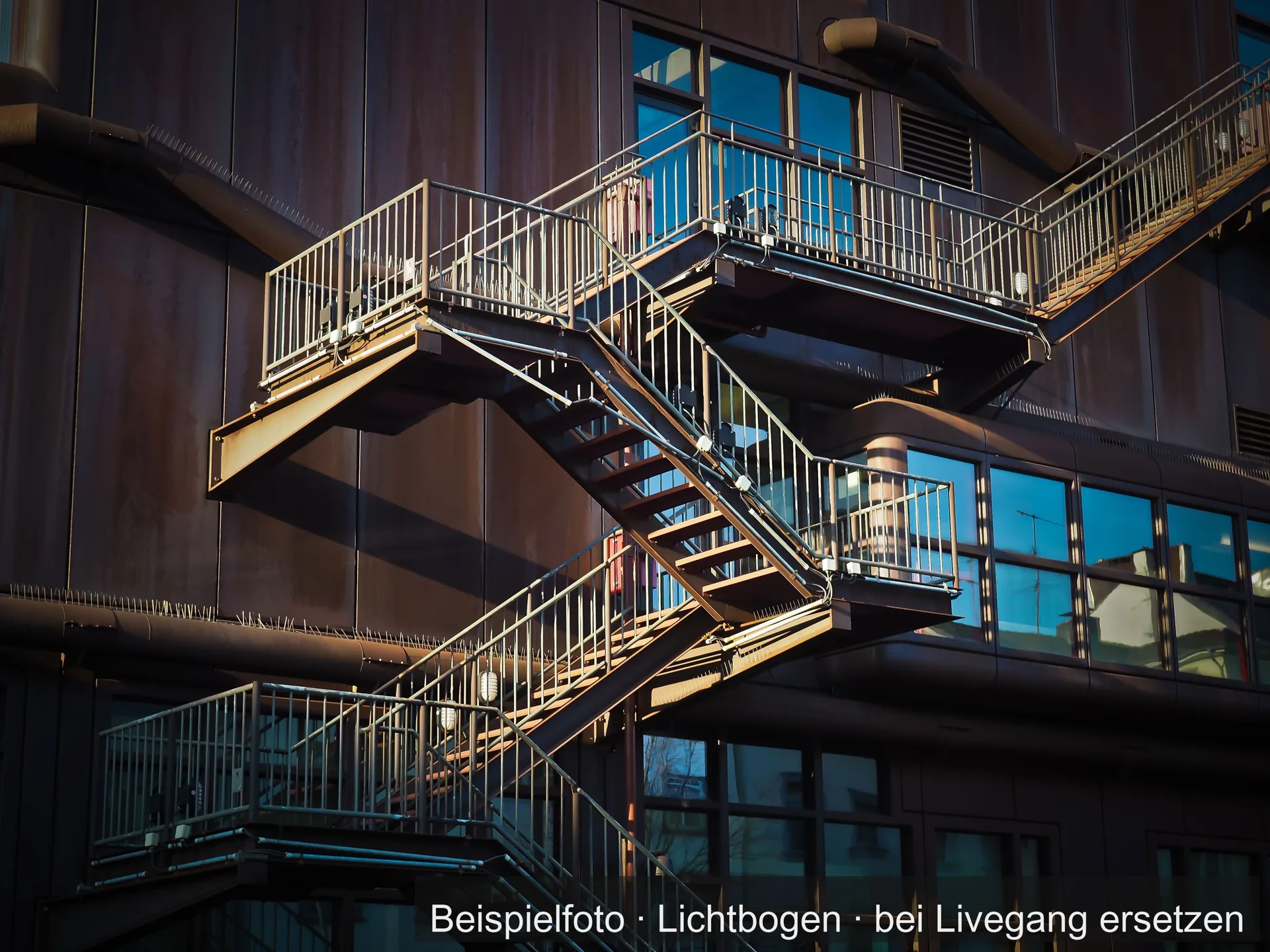 Außen liegende Stahltreppe mit Podesten und Geländern über mehrere Geschosse an der dunklen Fassade eines Gebäudes.