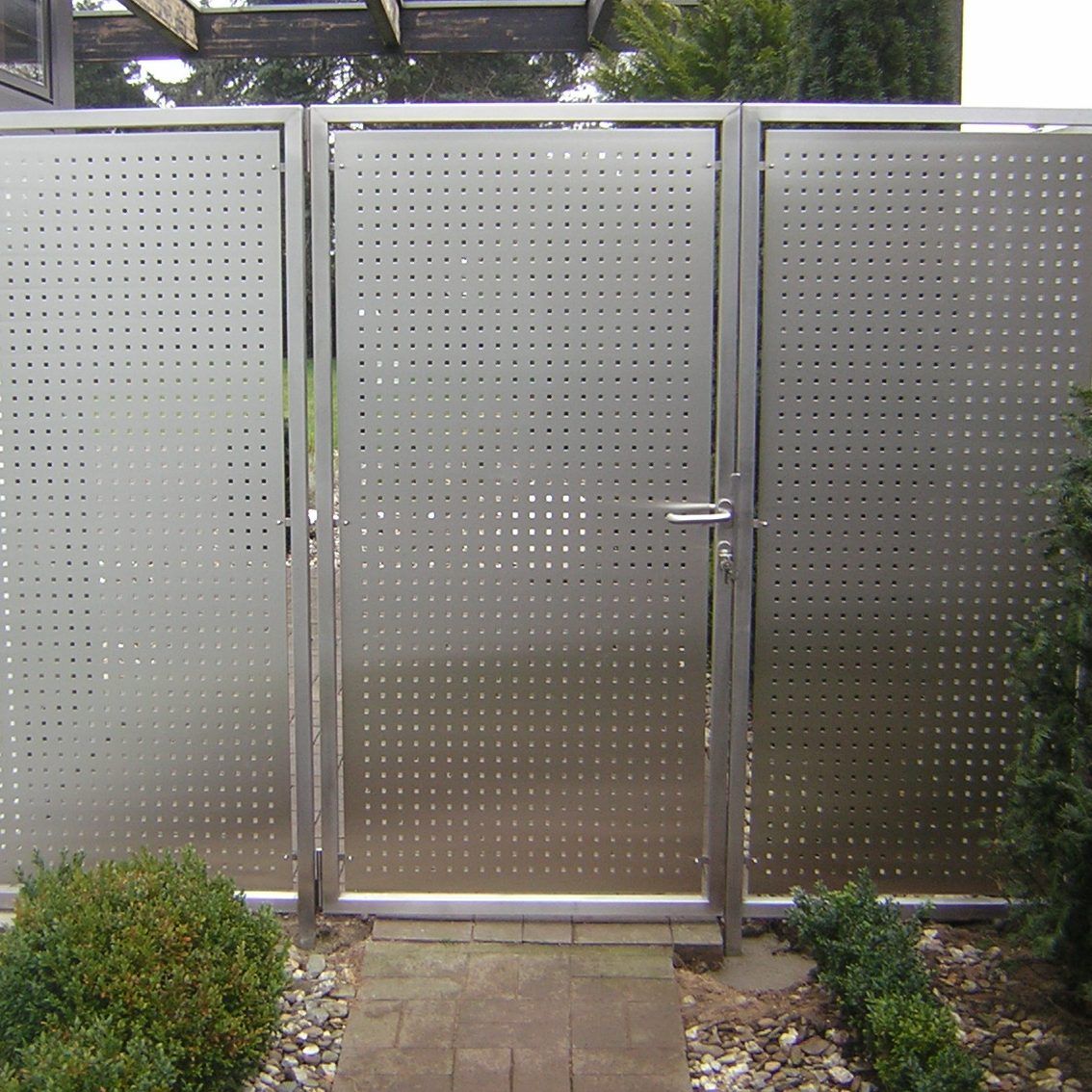 Zaunanlage aus Edelstahl 1.4301, 2 m hoch, mit einer Gartentür zwischen zwei feststehenden Feldern. Die Füllung ist Lochblech mit quadratischen Löchern von 10 × 10 mm als Sichtschutz; davor liegt ein gepflasterter Weg mit Buchs.