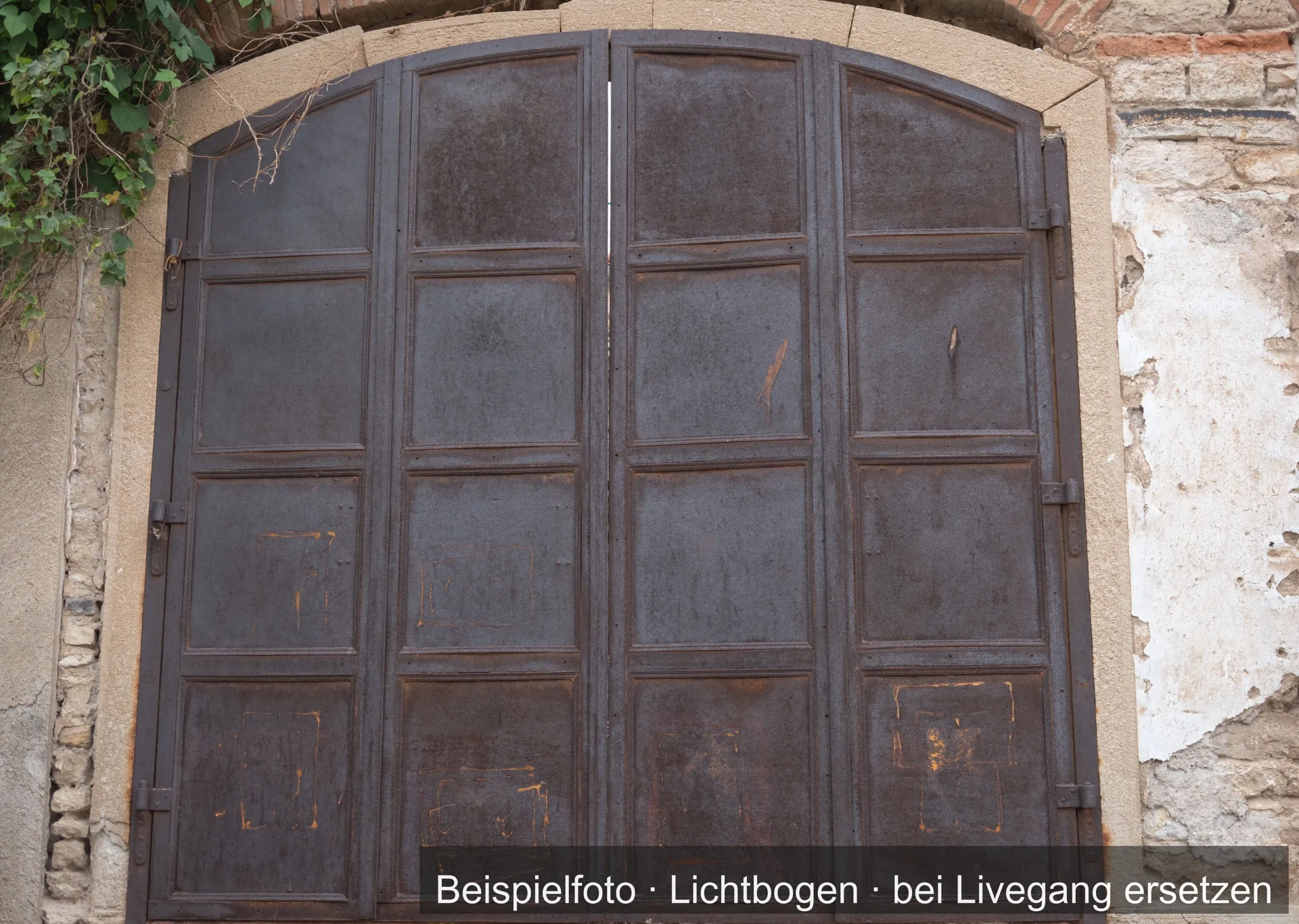 Zweiflügeliges Metalltor mit flachem Bogen und Kassettenfeldern in einer alten Mauer, die Oberfläche teils angerostet.