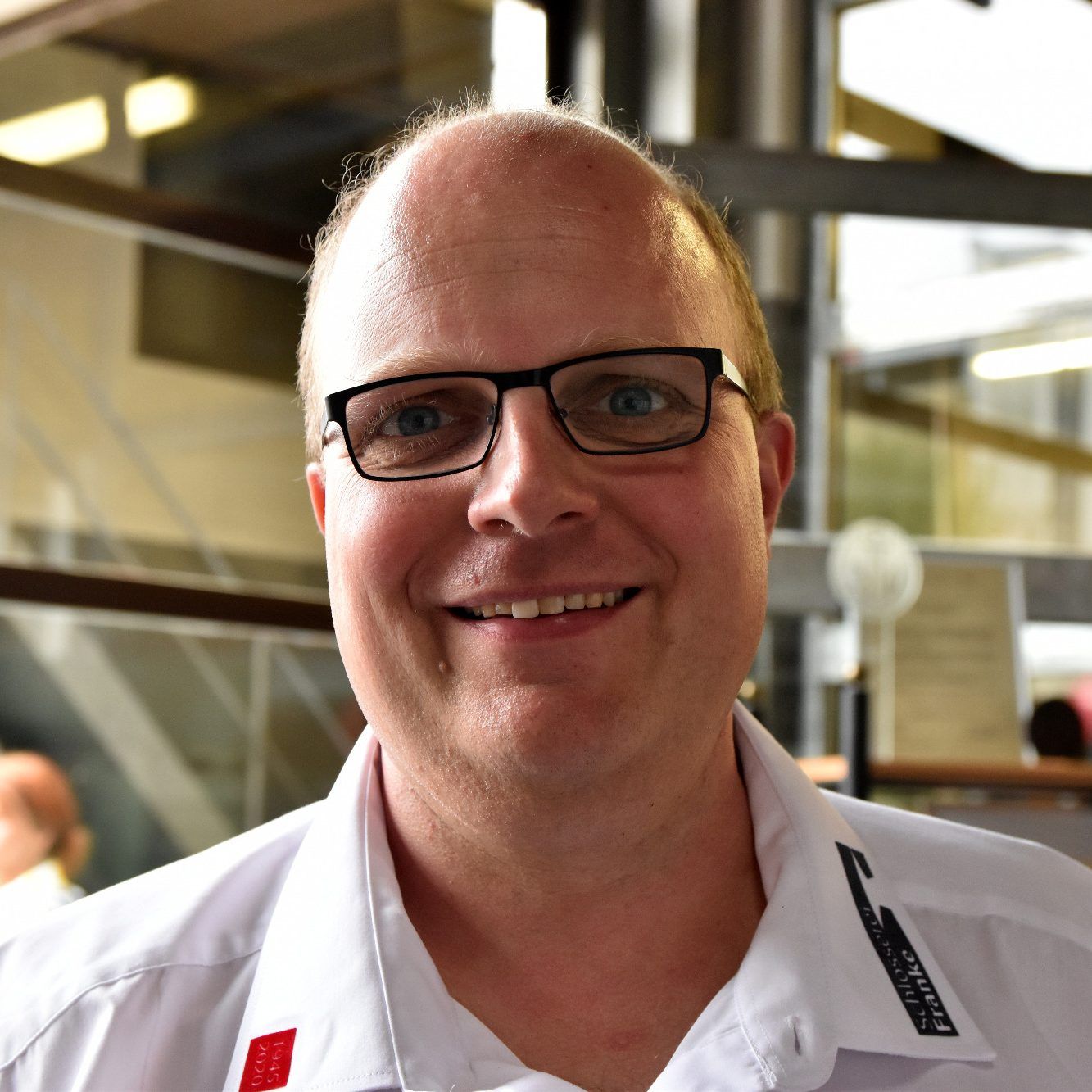 Porträt eines lächelnden Mannes mit Brille im weißen Firmenhemd, dahinter unscharf die Werkhalle.