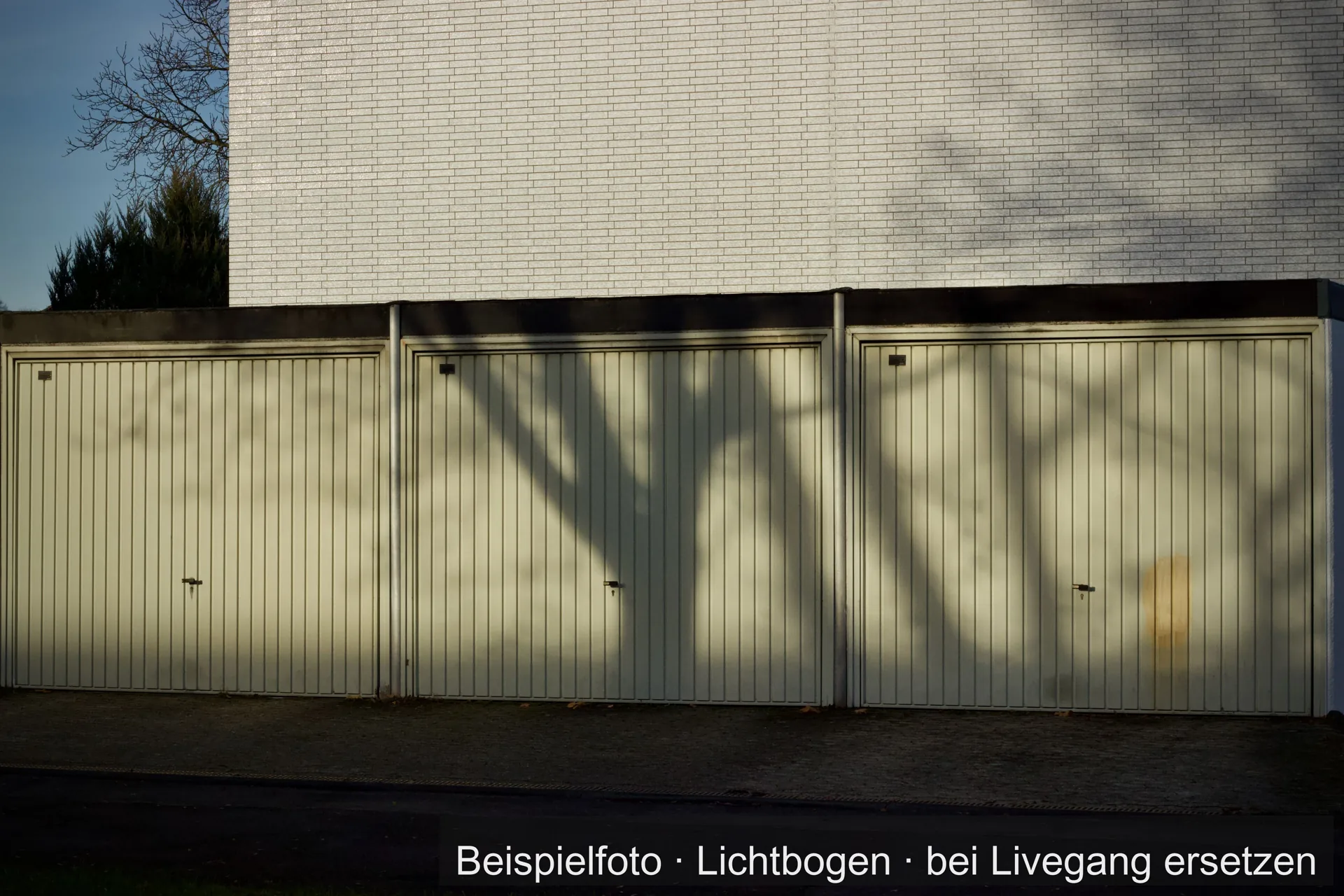 Drei helle Garagentore mit senkrechten Rillen in einer Garagenzeile vor einer weißen Klinkerwand, auf den Toren der Schatten eines Baums.