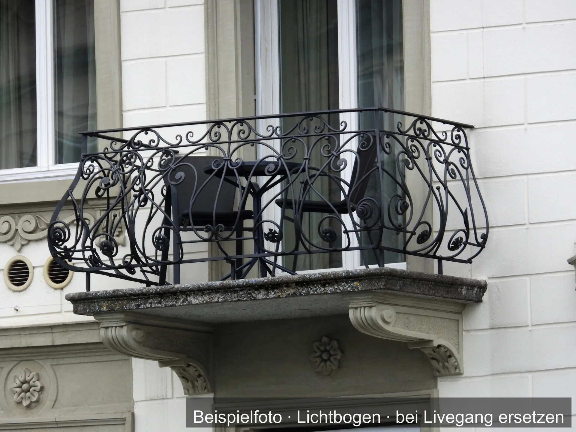 Balkon an einem hellen Altbau mit einem geschwungenen Geländer aus dunklem Metall mit Spiralornamenten; auf dem Balkon stehen ein Tisch und zwei Stühle.