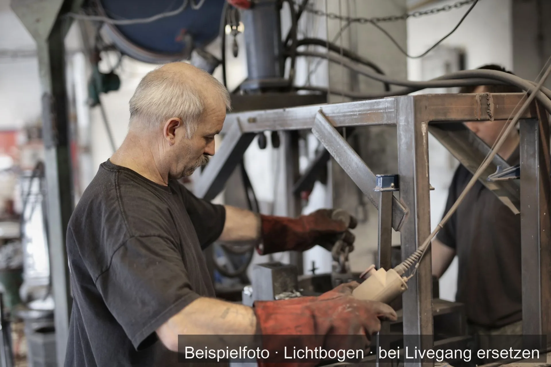 Zwei Metallbauer mit Schweißhandschuhen richten in einer hellen Werkstatt ein Gestell aus Stahl am Kettenzug aus.